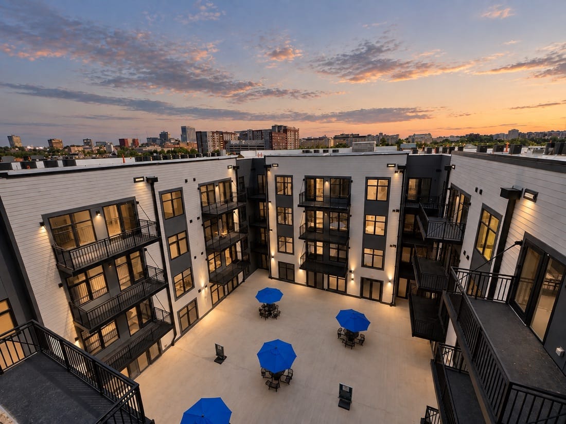 Modern apartment courtyard at dusk with blue umbrellas, lit windows, and city skyline at sunset.